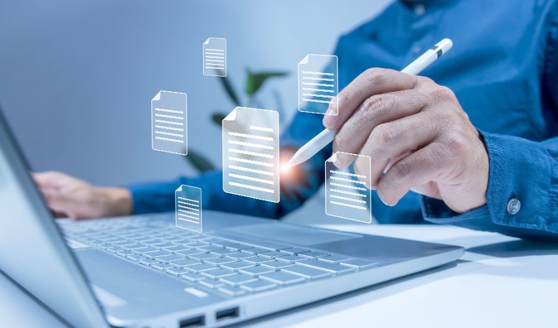 Eine Person tippt auf einem Laptop und auf davor eingeblendete Dokumenten-Icons. Zu sehen sind nur die Arme der Person.