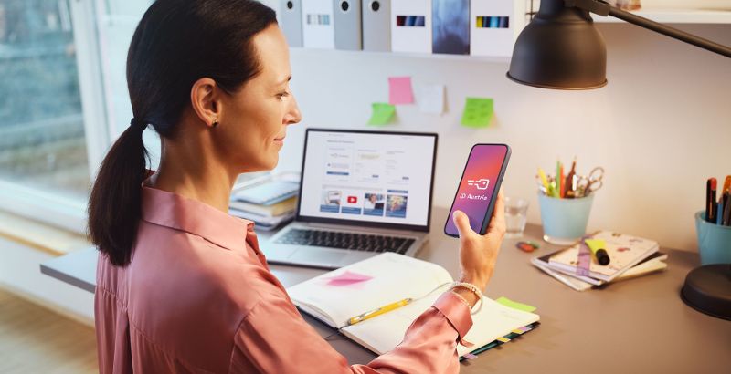 Eine Frau sitzt am Schreibtisch und arbeitet mit Laptop und Mobiltelefon, das sie in der Hand hält. Auf diesem ist die ID Austria zu sehen, die sie für die Anmeldung in den eServices der österreichischen Verwaltung verwendet.
